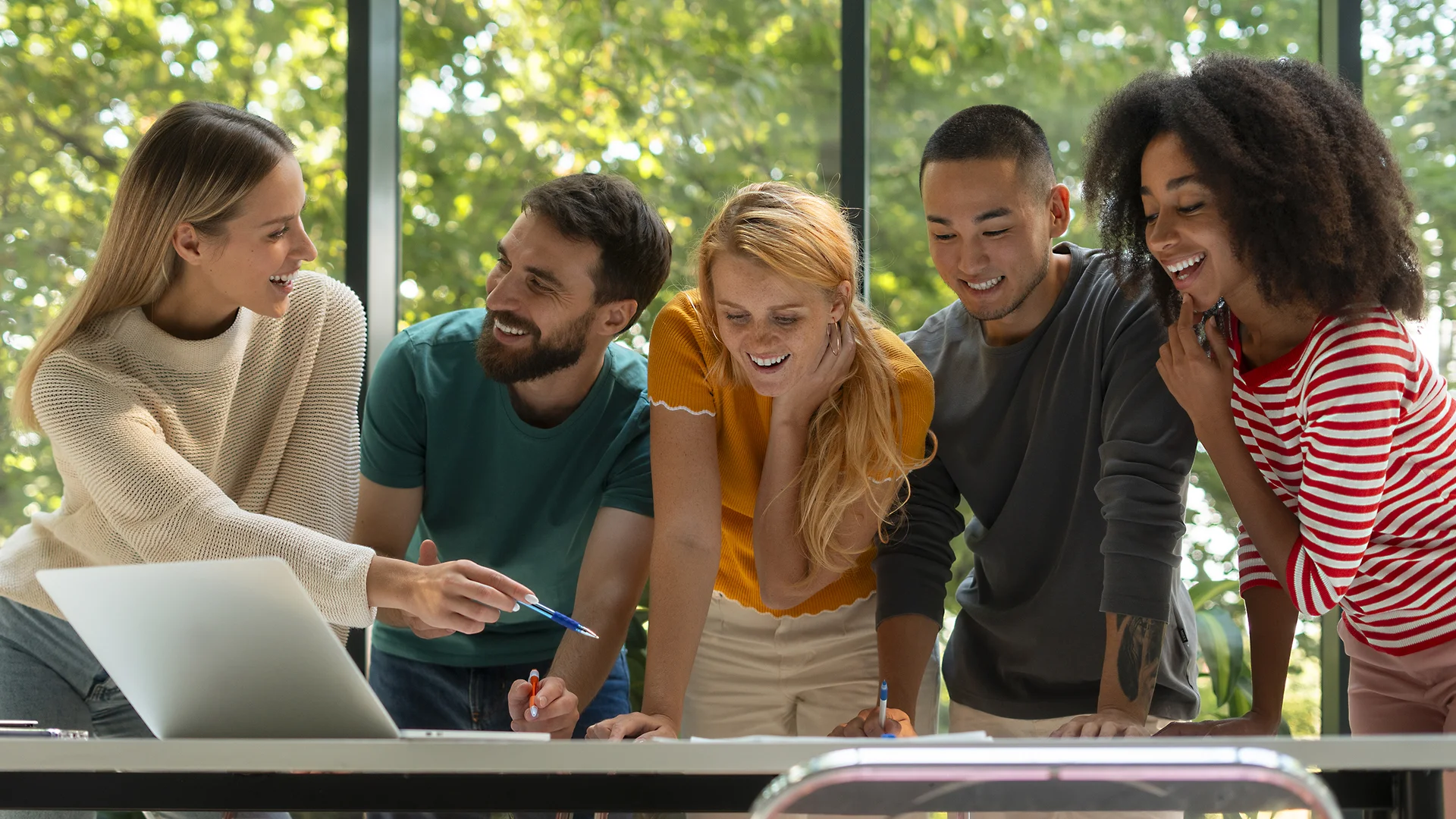 Grup de joves treballant junts al voltant d’una taula, somrient i col·laborant davant d’un portàtil en un espai lluminós.
